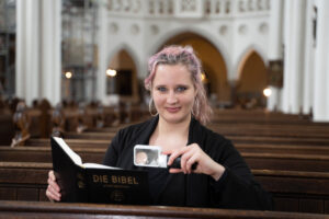 Eine Frau Mitte 20 sitzt in einer Kirchenbank und lächelt in die Kamera, in einer Hand eine Leuchtlupe, in der anderen eine Bibel. Sie hat langes, dunkelblondes Haar mit blonden und rötlichen Strähnchen und trägt große Ohrringe.