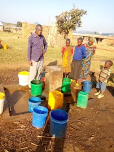 Mehrere Dorfbewohner stehen um den Brunnen herum. Vor ihren Füßen stehen mit Wasser gefüllte Behälter.